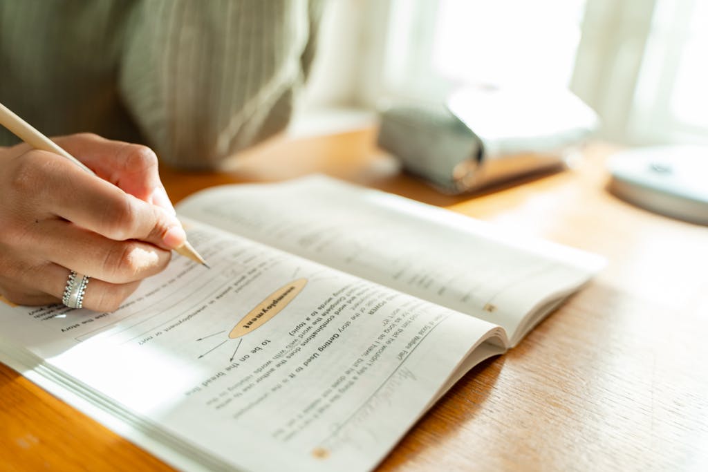 A student takes notes while studying at a sunlit desk with an open textbook.